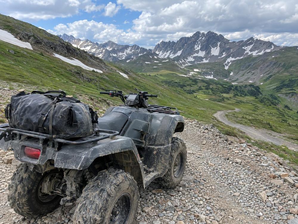Muddy ATV with a dry bag on a rocky switchback above treeline with snowy peaks behind