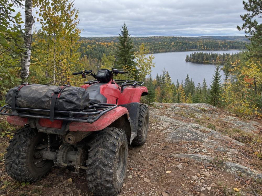 Red ATV with a strapped dry bag parked on a rocky trail above a forest lake