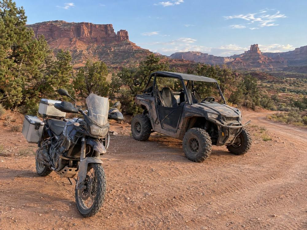 Adventure motorcycle and side-by-side parked together on a red dirt two-track below sandstone mesas
