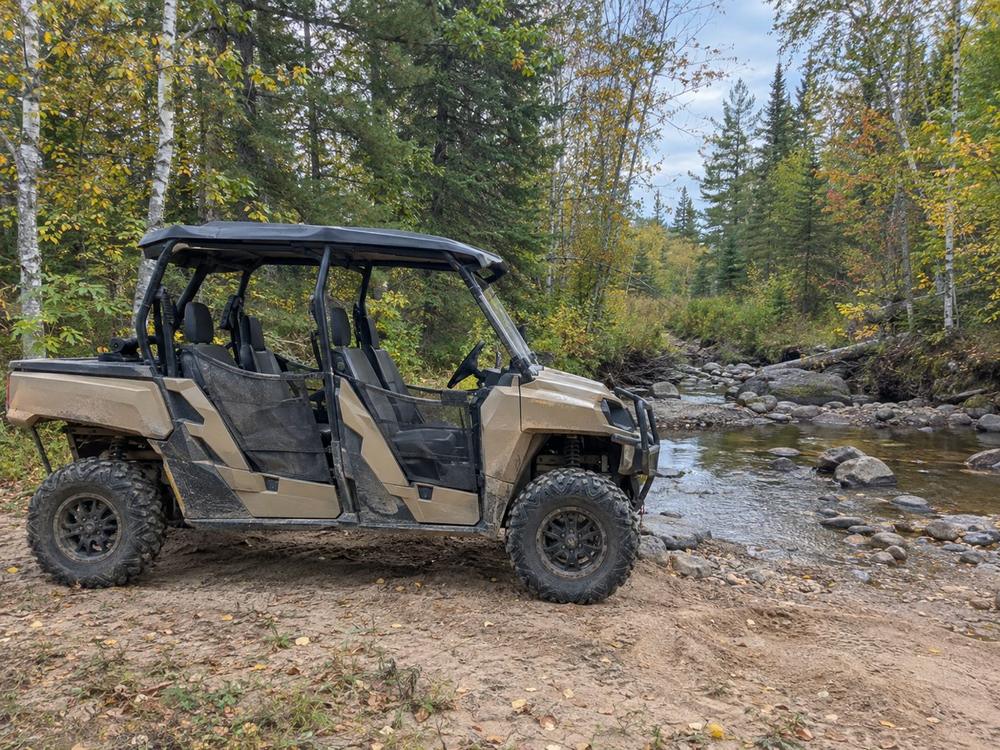 Tan four-seat side-by-side stopped beside a rocky creek crossing in the woods